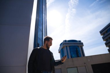 Sakallı, vücutlu ve güneş gözlüklü yakışıklı genç adam cam bir binanın önünde cep telefonundan e-posta ve sosyal ağlarını kontrol ediyor. Yeni teknoloji ve telefon kavramı.