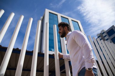 Sakallı, vücudu yontulmuş ve güneş gözlüklü yakışıklı genç adam beyaz çitlere tutunuyor. Fotoğraf aşağıdan çekildi ve arka planda mavi gökyüzünü görebilirsiniz..