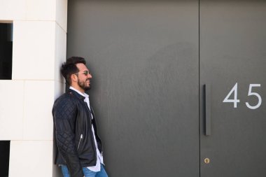 Sakallı, vücudu yontulmuş ve güneş gözlüklü yakışıklı genç adam portre için farklı pozlar verirken garajının gri kapısında duruyor. Farklı ifadeleri kavra.