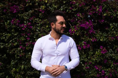 Sakallı, yontulmuş vücutlu yakışıklı bir genç adam portresi. Arka planda yeşil ve mor tırmanma bitkileri var. Fotoğraflar için farklı yerlere bakıyor..