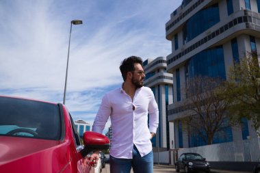 Yakışıklı bir genç adam, kırmızı spor arabasının yanında heykel gibi bir vücudu var. Adam zengin ve modern kıyafetler giymiş. Spor araba.
