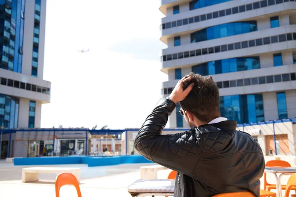 young and handsome man with beard, sculptural body, is sitting in a bar while touching his hair. In the horizon you can see a plane in the sky. Hospitality and catering concept, restaurants and travel