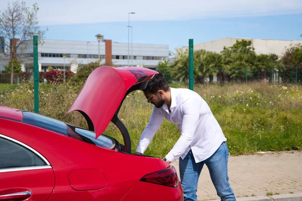 Young, handsome man opens the boot of his red sports car. The man is wealthy. High standard of living and well positioned financially.