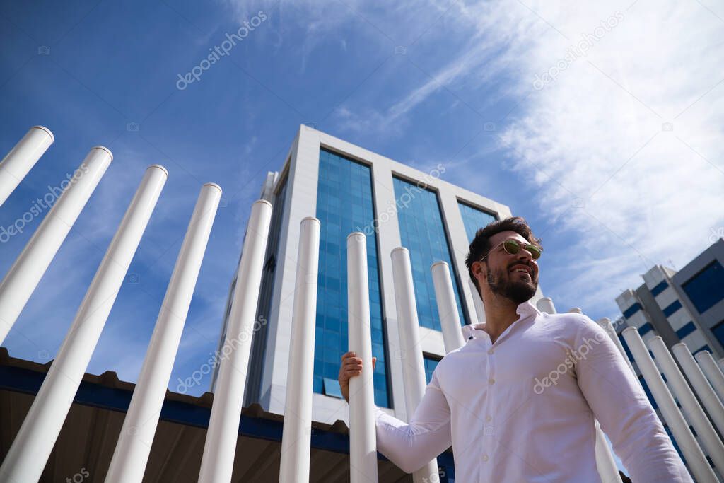handsome young man with beard, sculpted body and sunglasses is clinging to a white picket fence. The photo is taken from below and you can see the blue sky in the background.