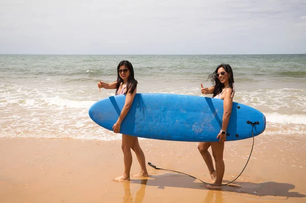 Two young and beautiful women with a surfboard on the shore of the beach. The women are enjoying their trip to the beach paradise. Holidays and travels.