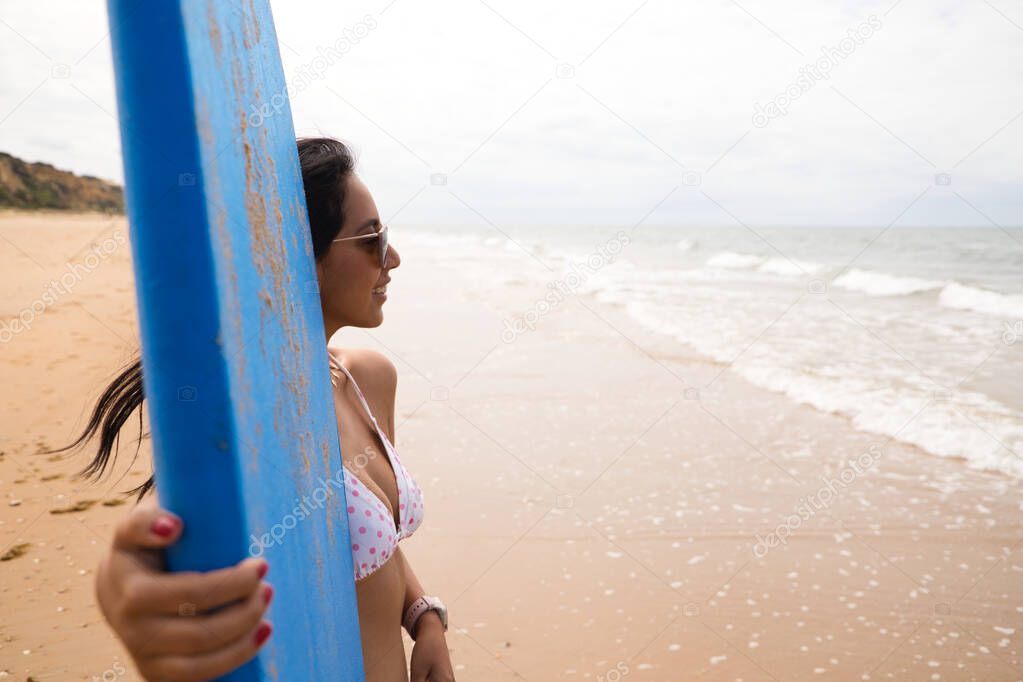 Beautiful young latin woman in bikini and with blue surfboard. The woman is on the beach. Holiday and summer concept.