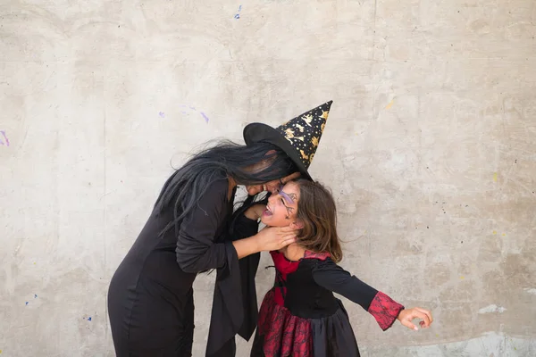 Happy halloween. Young beautiful woman and girl dressed as witches on grey background. The woman is giving kisses to the girl on her face on the day of halloween party. Trick or treat.