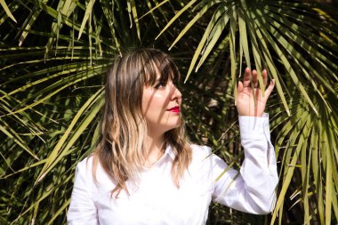 Güzel, sarışın, genç bir kadın parkta yeşil palmiye ağaçlarının önünde farklı ifadeler takınarak eğleniyor. Tatil ve boş zaman kavramı. Çeşitli ifadeler.