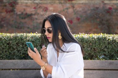 Avrupa 'da tatilde olan genç ve güzel bir Güney Amerikalı kadın cep telefonunu ayna olarak kullanarak bir bankta dudaklarını boyuyor..