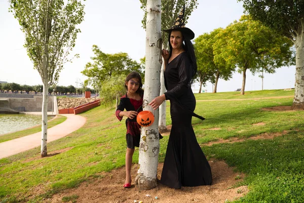 Happy halloween. Young beautiful woman and girl dressed as witches next to a tree and with a pumpkin to collect candy and sweets. Trick or treat. 31st October.