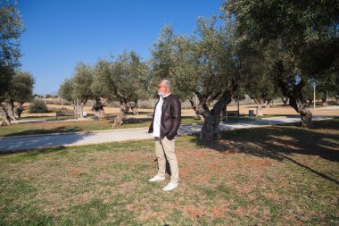 Sakallı ve gri saçlı yakışıklı olgun bir adam parkta yürüyor. Adam bej pantolon, beyaz gömlek ve deri ceket giyiyor. Yaşlılık kavramı