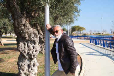 Sakallı ve gri saçlı yakışıklı olgun bir adam parkta dalgın dalgın bir şekilde sokak lambasına yaslanıyor. Adam bej pantolon, beyaz gömlek ve deri ceket giyiyor. Yaşlılık kavramı