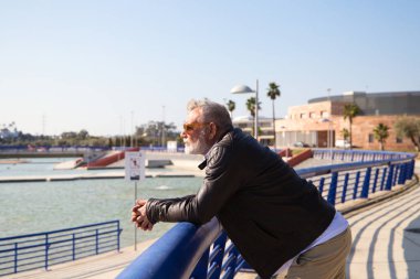 Sakallı ve gri saçlı yakışıklı olgun bir adam parkta yürüyor. Adam bej pantolon, beyaz gömlek ve deri ceket giyiyor. Yaşlılık kavramı