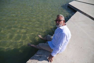 Sakallı ve gri saçlı yakışıklı bir adam yerde oturuyor ve ayakları parkta bir gölde sıkıştı. Adam bej pantolon ve beyaz gömlek giyiyordu. Yaşlılık, seyahat ve turizm kavramları