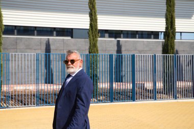 Handsome, mature, grey-haired man with beard, jacket and tie and sunglasses is on the university campus where he works as a university lecturer. Concept professor, university rector.