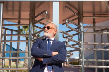 Handsome, mature, grey-haired man with beard, jacket and tie and sunglasses at the entrance to the office building where he works as chief accountant and executive. The man with his arms crossed.