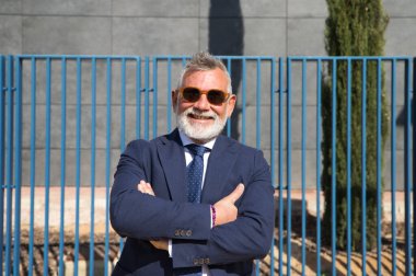 Yakışıklı, olgun, gri saçlı, sakallı, ceketli, kravatlı ve güneş gözlüklü bir adam. Baş muhasebeci ve yönetici olarak çalıştığı ofis binasının girişinde. Olgun iş adamı konsepti