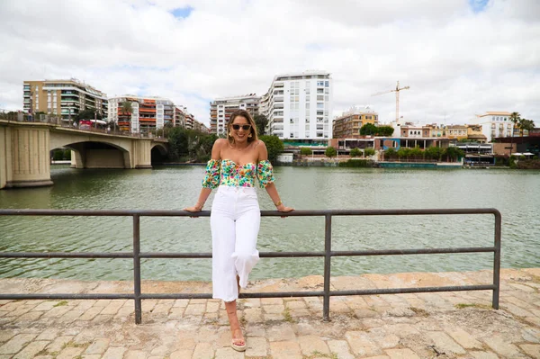 Young, pretty blonde woman with sunglasses leaning on the railing of the river. The woman is happy and smiling. In the background you can see the river in seville.