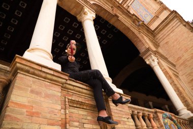 Esmer saçlı ve dans kıyafetleri giymiş güzel genç flamenko dansçısı iki beyaz mermer sütun arasında oturuyor ve kırmızı yelpaze ile flamenko dansı yapıyor..