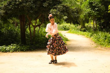 A beautiful teenage flamenco dancer dressed in a flowery skirt with ruffles and flowers in her hair is dancing in the park with a lot of art. Flamenco concept cultural heritage of humanity.