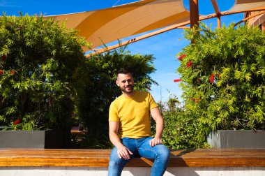 Mavi gözlü, sakallı ve sarı tişörtlü yakışıklı genç bir adam yeşil bitkilerin yanındaki bankta oturuyor. Adam mutlu ve gülümsüyor..