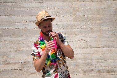 Hawaii çiçekli gömlek, şapka ve renkli çiçek kolyesi giyen yakışıklı genç adam, içinde meşrubat olan bir bardaktan bir kamışdan su içiyor. Tatil ve seyahat kavramı.