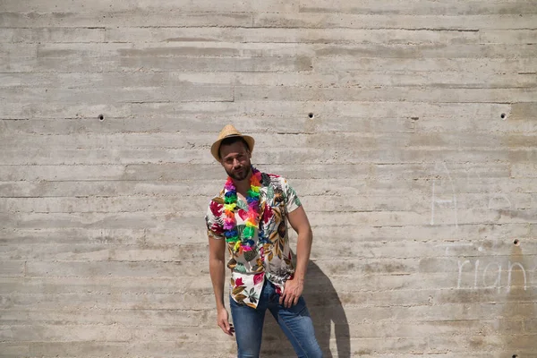 一个英俊的年轻人 穿着一件有夏威夷花的衬衫 头戴一顶帽子 戴着一条有五颜六色花的项链 这个人很快乐 背景灰墙壁 旅行和假日概念 — 图库照片