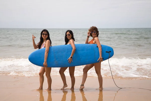 Three young and beautiful women on a surfboard on the shore of the beach. The women are enjoying their trip to the beach paradise. Holidays and travels.