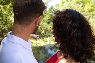 A handsome, young Latin man and woman are in the forest and point to a lake. The couple are engaged and are spending a day in the countryside.
