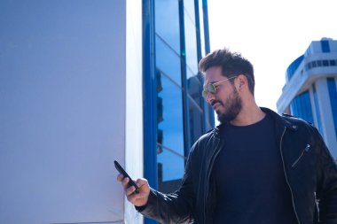 Sakallı, vücutlu ve güneş gözlüklü yakışıklı genç adam cam bir binanın önünde cep telefonundan e-posta ve sosyal ağlarını kontrol ediyor. Yeni teknoloji ve telefon kavramı.