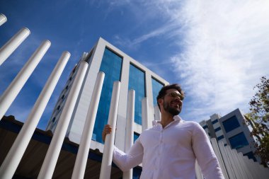 Sakallı, vücudu yontulmuş ve güneş gözlüklü yakışıklı genç adam beyaz çitlere tutunuyor. Fotoğraf aşağıdan çekildi ve arka planda mavi gökyüzünü görebilirsiniz..
