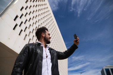 Sakallı, heykel vücutlu yakışıklı genç adam sokakta cep telefonuyla selfie çekiyor..