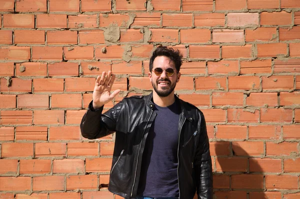 Genç Latin bir adam, esmer, sakallı ve el kaldırarak vücut bulmuş. Arka planda kırmızı tuğla duvar var. Merhaba de, güle güle, selamla. İnsan ilişkileri ve duyguları.