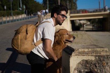 A handsome young man with his brown golden retriever dog leaning on the railing overlooking the river. They are on holiday in seville, spain. Concept pets, animals, dogs, pet love, travel.