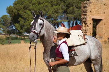 Young and beautiful Spanish woman with her thoroughbred horse. The woman is wearing a riding uniform and a typical Spanish hat. They are next to the stable in the countryside. Concept equines.