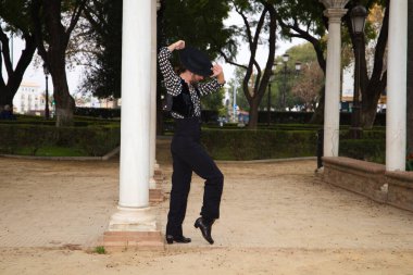 Çingene adam uzun saçlı ve sakallı flamenko dansı yapıyor mermer sütunların yanındaki parkta. Elinde siyah bir şapka tutuyor ve tutkuyla farklı duruşlar sergiliyor. Flamenko dansı.