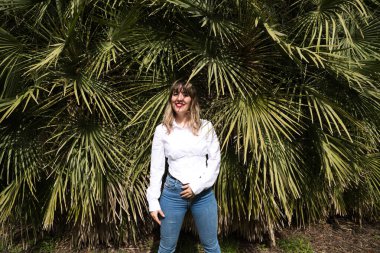 Güzel, sarışın, genç bir kadın parkta yeşil palmiye ağaçlarının önünde farklı ifadeler takınarak eğleniyor. Tatil ve boş zaman kavramı. Çeşitli ifadeler.