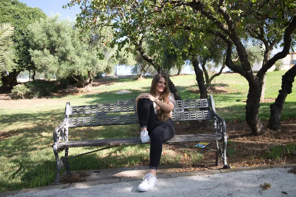 ブロンドの髪と青い目をした美しい若い女性が公園のベンチに座って モデルのような写真のポーズをとっています 身体の体位や表情が違うのが嬉しい — ストック写真