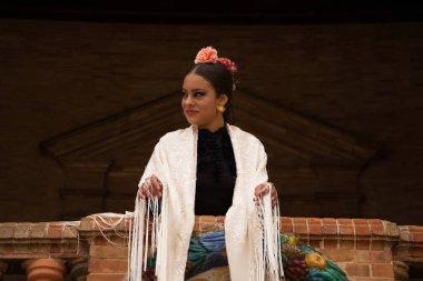 A beautiful teenage flamenco dancer with brown hair stands on a balcony wearing a shawl embroidered with fringes and performs flamenco dance postures. World Cultural Heritage.
