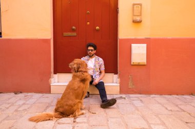 Genç, yakışıklı İspanyol bir adam, kahverengi köpeğiyle, bir basamakta dinleniyorlar, arka planda kırmızı bir kapı. Evcil hayvanlar, hayvanlar, köpekler, evcil hayvanlar, Golden Retre