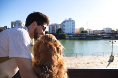 A handsome young man with his brown golden retriever dog leaning on the railing overlooking the river. They are on holiday in seville, spain. Concept pets, animals, dogs, pet love, travel.