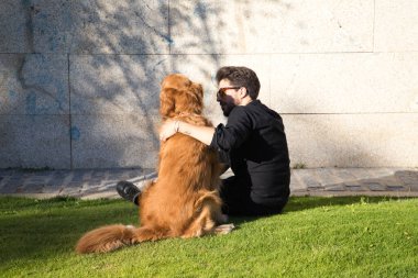 Sakallı ve gözlüklü yakışıklı genç adam çimenlerde oturmuş köpeğiyle güneşleniyor. Evcil hayvanlar, hayvanlar, köpekler, evcil hayvanlar, seyahat.