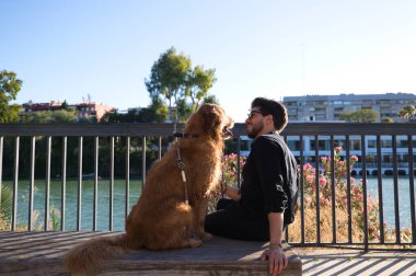 A handsome young man sitting with his golden retriever dog on a bench. In the background you can see the river and against the sunlight in the evening. Concept pets, animals, dogs, pet love.