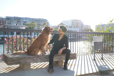A handsome young man sitting with his golden retriever dog on a bench. In the background you can see the river and against the sunlight in the evening. Concept pets, animals, dogs, pet love.