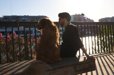 A handsome young man sitting with his golden retriever dog on a bench. In the background you can see the river and against the sunlight in the evening. Concept pets, animals, dogs, pet love.