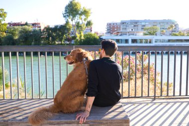 A handsome young man sitting with his golden retriever dog on a bench. In the background you can see the river and against the sunlight in the evening. Concept pets, animals, dogs, pet love.