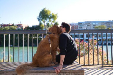 A handsome young man sitting with his golden retriever dog on a bench while giving him a kiss. In the background you can see the river and against the sunlight in the evening. Concept pets, animals.