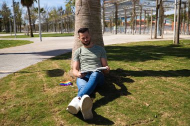 Yakışıklı genç homoseksüel adam parkta çimenlerde oturmuş kitap okuyor, yanında da eşcinsel gurur bayrağı var. Konsept okuma, eğitim, öğretim, boş zaman ve boş zaman.