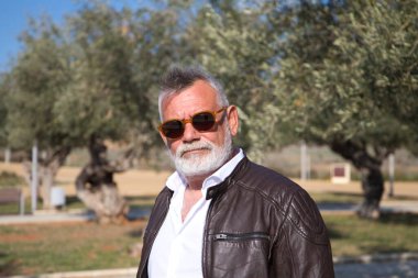 Sakallı ve gri saçlı yakışıklı olgun bir adam parkta yürüyor. Adam bej pantolon, beyaz gömlek ve deri ceket giyiyor. Yaşlılık kavramı