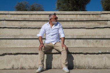 Sakallı ve gri saçlı yakışıklı olgun bir adam açık hava konferans salonunun merdivenlerinde oturuyor. Adam farklı yerlere bakıyor. Kıdemli konsept, seyahat ve turizm.